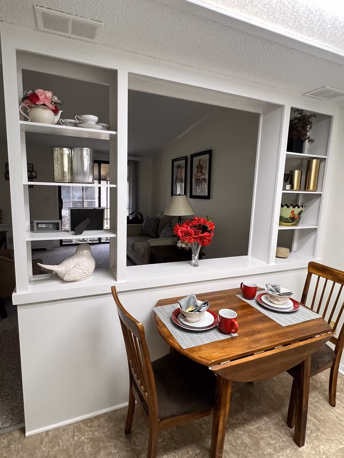 A small wooden dining table set for two with red mugs, bowls, and plates on placemats. The table is positioned against a half wall with built-in shelves holding decorative items like a white bird figurine, a teapot with flowers, and gold containers. Through the opening in the wall, a living room with a sofa, lamp, and framed pictures on the wall is visible.
