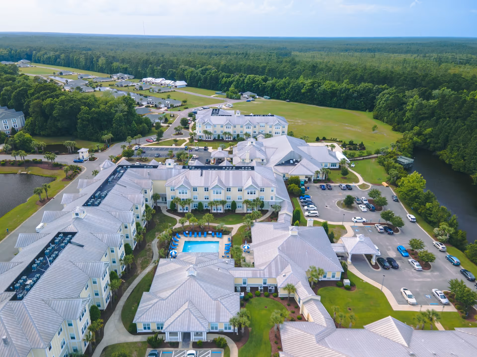 Aerial view of a large senior living facility named Brightwater, featuring multiple connected buildings with light-colored roofs, a central outdoor swimming pool with blue lounge chairs, surrounding parking lots with cars, green lawns, trees, and a nearby pond.