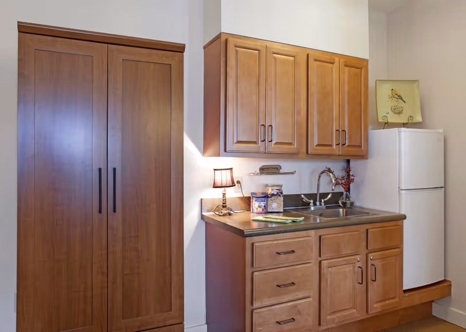 A small kitchen area with wooden cabinets, a countertop with a sink, a small lamp, and a white refrigerator. There is a decorative plate with a bird on top of the refrigerator and a wooden pantry cabinet to the left.
