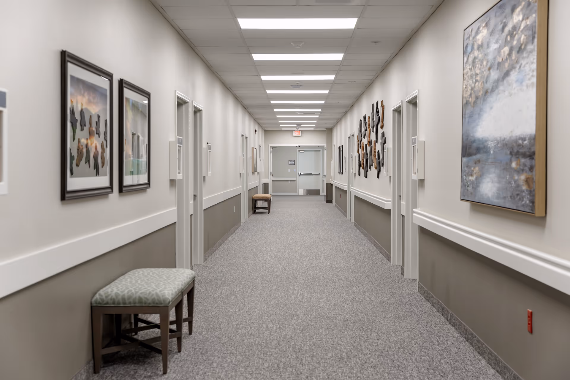 A long, well-lit hallway in a senior living facility with beige walls, gray carpet, and multiple closed doors on both sides. The walls are decorated with framed artwork and sculptures, and there are cushioned benches placed along the hallway. The ceiling has recessed lighting panels, and an exit door is visible at the far end.