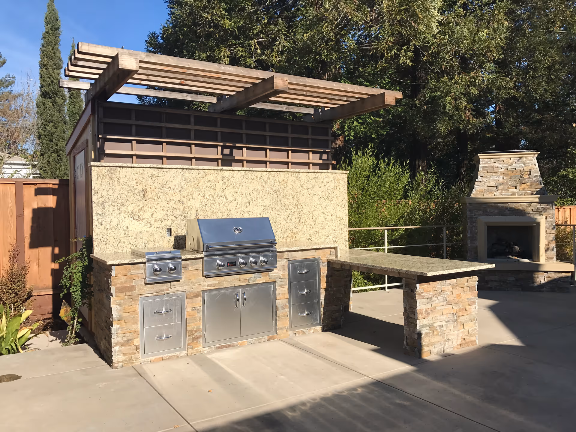 Outdoor patio area with a built-in stone and stainless steel barbecue grill, a stone countertop table, and a stone fireplace surrounded by trees and a wooden fence under a clear blue sky.