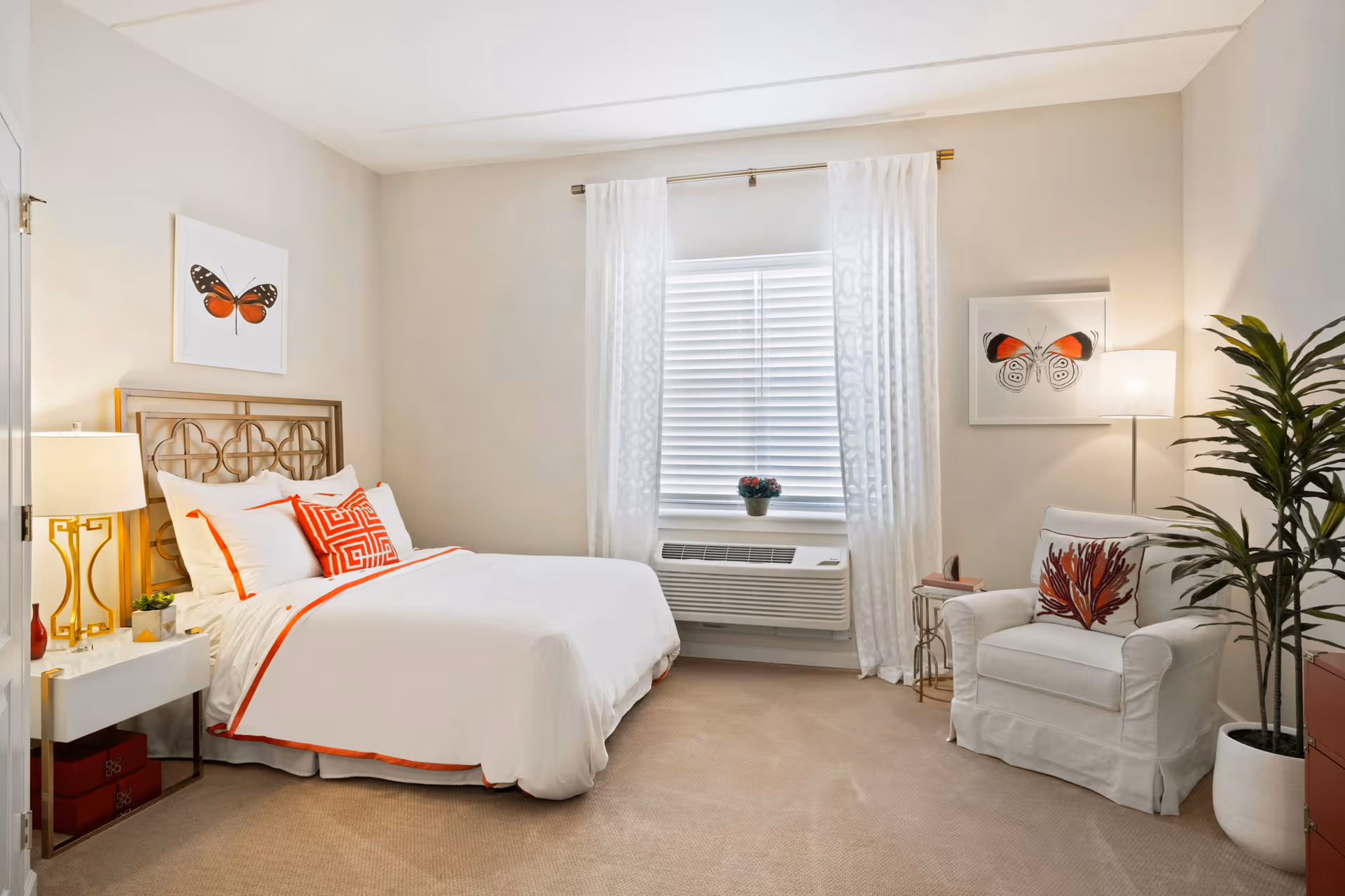 Bright furnished bedroom with a bed, bedside table and lamp, armchair, potted plant, and butterfly artwork by a curtained window.