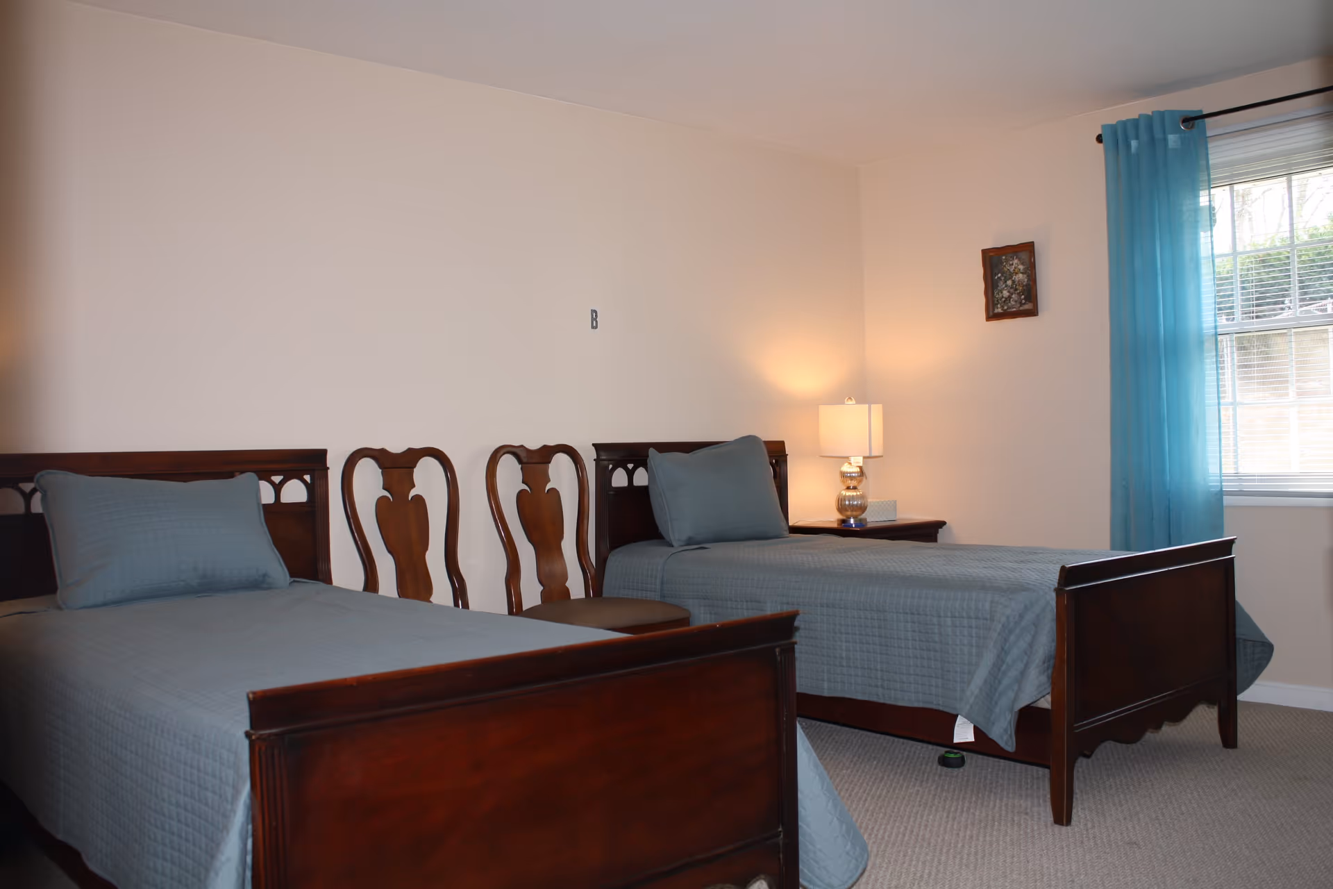 A bedroom with two wooden twin beds covered with matching blue-gray quilts and pillows. Between the beds are two wooden chairs and a nightstand with a lamp and a box of tissues. A window with blue curtains is on the right wall, and a small framed picture hangs above the nightstand.