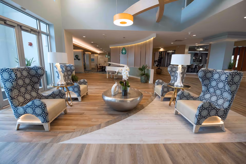 A modern and spacious senior living facility common area with four patterned armchairs arranged around a round metallic coffee table with a white orchid plant. Two side tables with white lamps are placed between the chairs. The room features wood flooring, large windows, and contemporary lighting fixtures, including a hanging pendant light. In the background, there is a white grand piano and additional seating areas.