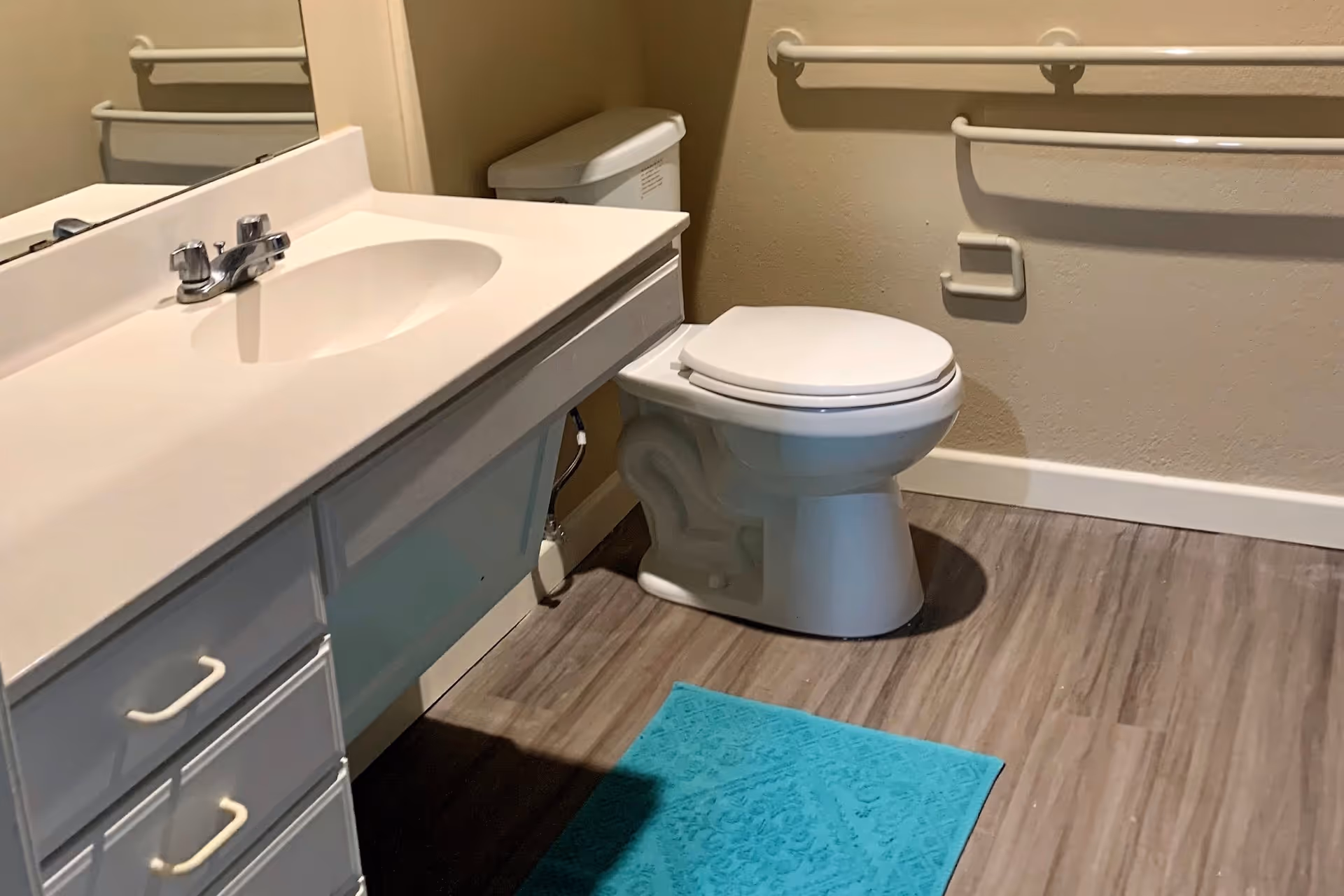 A bathroom with a white sink and countertop on the left, a white toilet with the lid down on the right, beige walls with grab bars installed, and a blue bath mat on the wood-patterned floor.