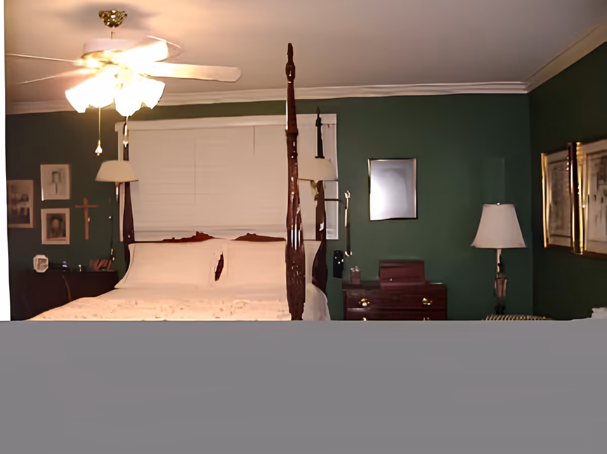 Four-poster bed with white bedding in a bedroom with green walls, ceiling fan, lamps, and dressers.