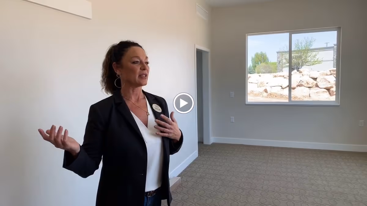 A woman in a black blazer and white blouse is standing in an empty room with beige patterned carpet and light-colored walls. She appears to be speaking or presenting. There is a window on the right side showing an outdoor view with rocks and trees.