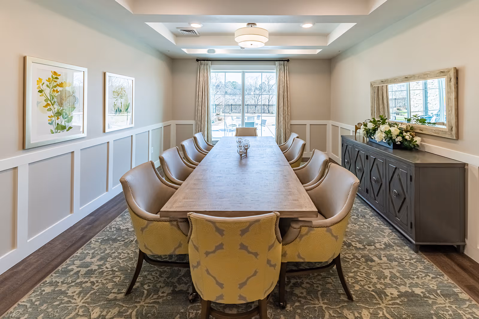 A bright and elegant dining room with a long wooden table surrounded by ten upholstered chairs. The room features light-colored walls with white wainscoting, two framed floral artworks on the left wall, a large window with curtains at the far end, and a sideboard with a decorative mirror and floral arrangement on the right. A patterned rug covers the floor beneath the table and chairs.