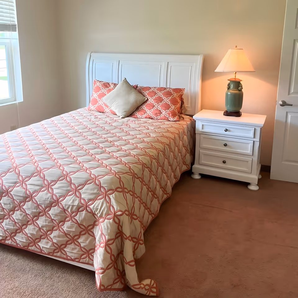 A bedroom with a white wooden bed frame and a bed made with a patterned quilt in shades of cream and coral. There are two coral patterned pillows and one beige pillow on the bed. Next to the bed is a white nightstand with three drawers and a green ceramic lamp with a white lampshade. A window with blinds is visible on the left side, and a closed white door is on the right.