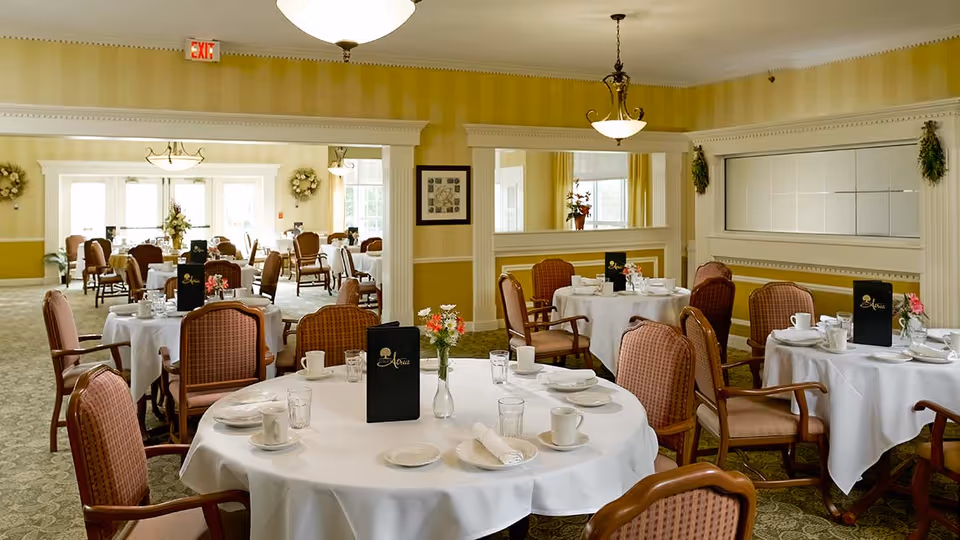 A bright and elegant dining room with round tables covered in white tablecloths, set with plates, cups, glasses, and napkins. The room features upholstered wooden chairs, yellow striped wallpaper, decorative wall moldings, and hanging light fixtures. Menus and small flower vases are placed on the tables.