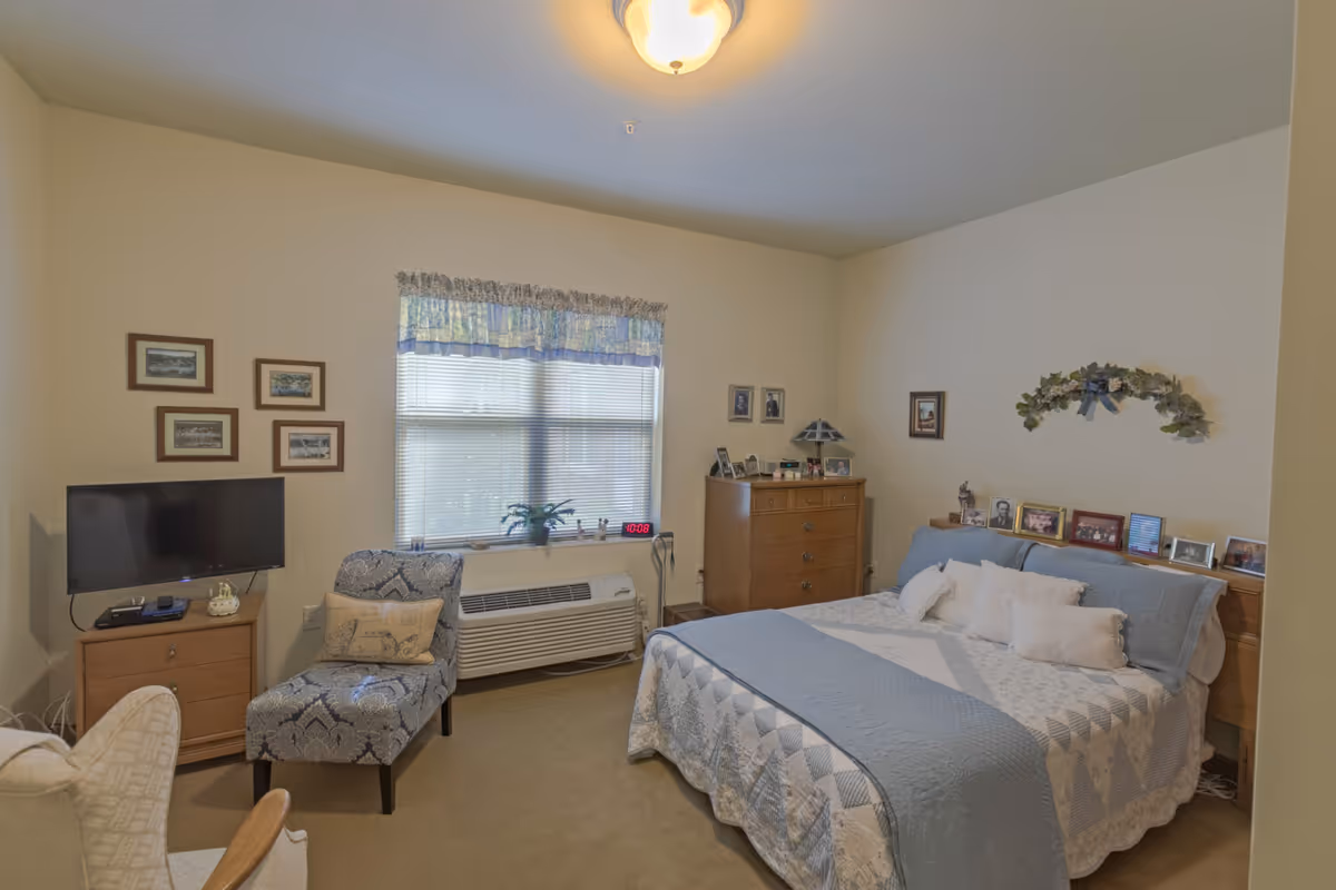 Cozy, well-lit bedroom with a blue-and-white quilted bed, wooden dressers, a TV, armchair, and a window with a valance.