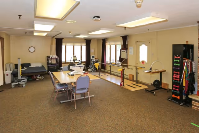 A rehabilitation room with exercise equipment including parallel bars for walking practice, a table with chairs, a hospital bed, and various physical therapy tools. The room has beige walls, carpeted floor, and large windows with dark curtains allowing natural light.