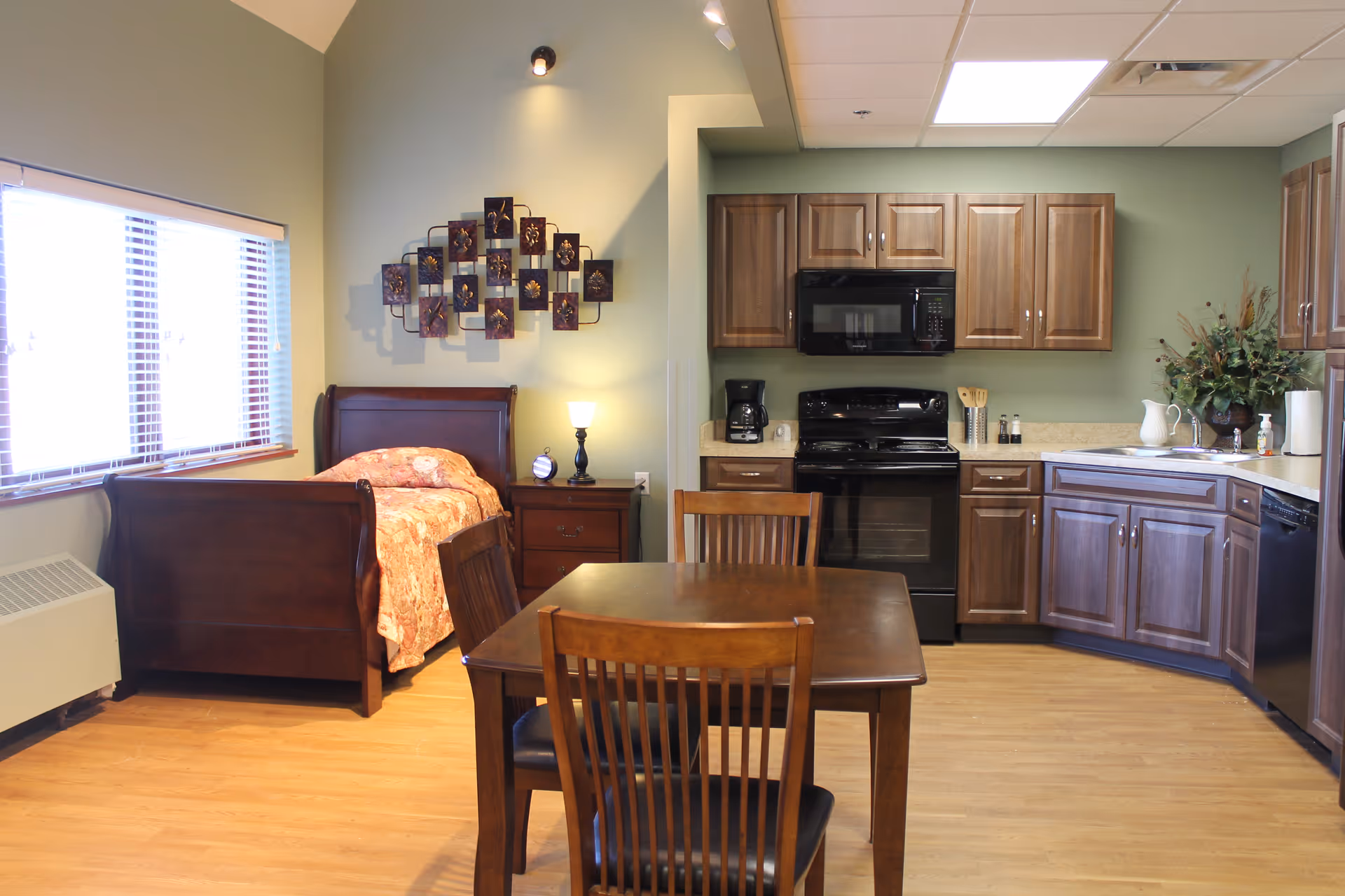A combined living space featuring a wooden single bed with a floral bedspread, a nightstand with a lamp and clock, a wooden dining table with chairs, and a kitchen area with wooden cabinets, a black stove, microwave, coffee maker, and a sink. The room has light green walls and a large window with blinds letting in natural light.