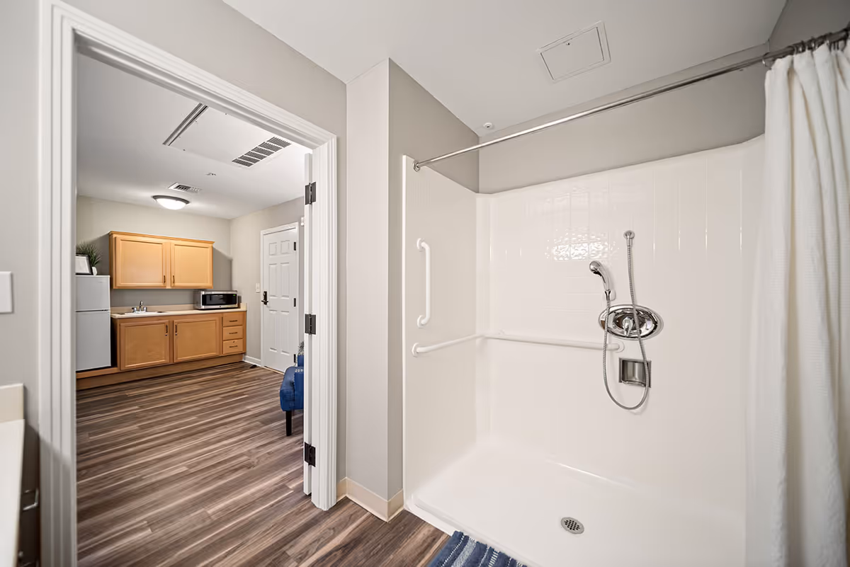 Interior view of a bathroom with a walk-in shower featuring grab bars and a handheld showerhead. The shower has a white curtain and white tiled walls. Through an open door, a kitchenette area with wooden cabinets, a small refrigerator, a microwave, and a sink is visible. The flooring is wood-style laminate throughout.