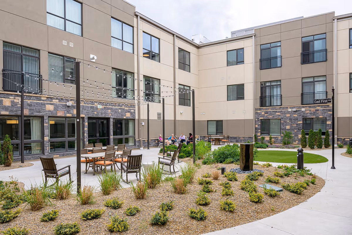 Outdoor courtyard area of Hilltop Reserve Senior Living with seating arrangements, string lights overhead, landscaped garden beds, and a group of people sitting and conversing near the building.