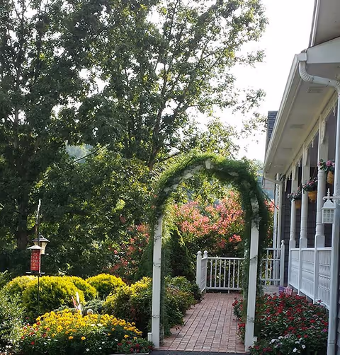 Brick walkway through a lush flower garden under a white arbor leading to the porch of a house.
