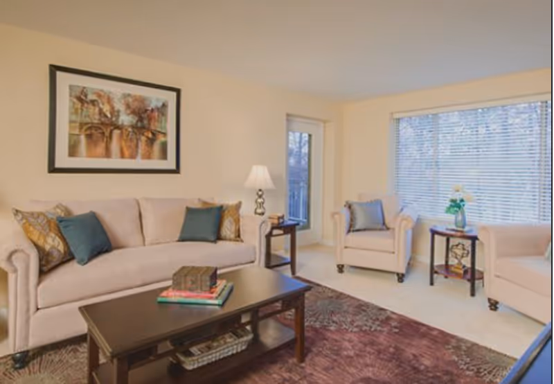 Bright living room with a sofa, two armchairs, a coffee table, side tables with a lamp and vase, and a large window with blinds.