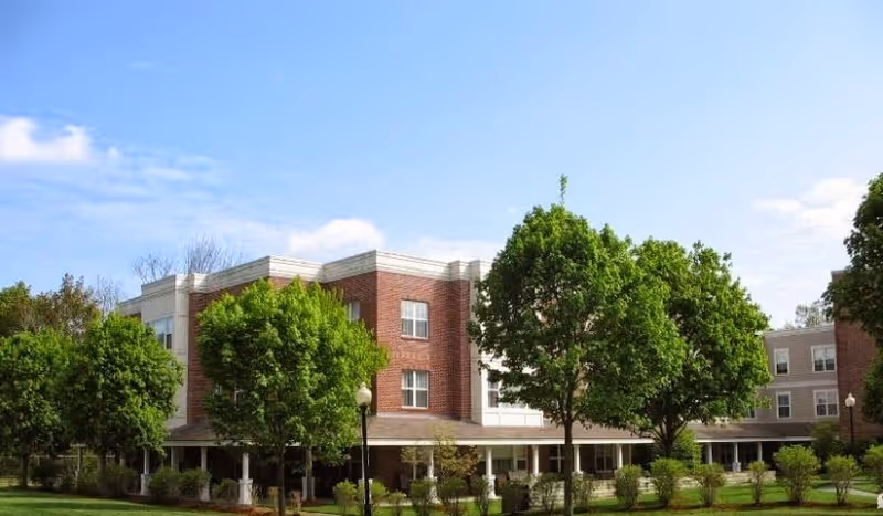 Exterior view of a multi-story senior living facility building with red brick and white accents, surrounded by green trees and a well-maintained lawn under a clear blue sky.