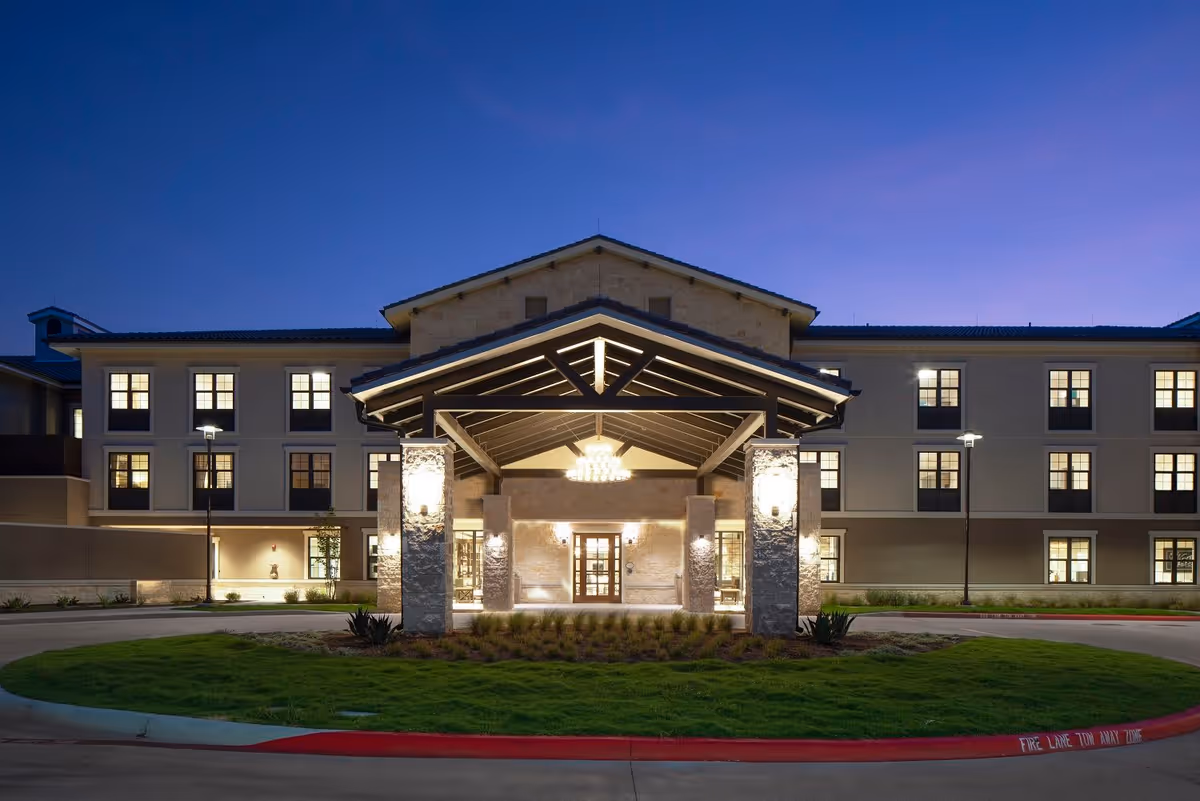 Exterior front view of Belmont Village Senior Living Lakeway building at dusk, featuring a well-lit entrance with stone pillars, a covered driveway, and multiple windows with lights on inside.