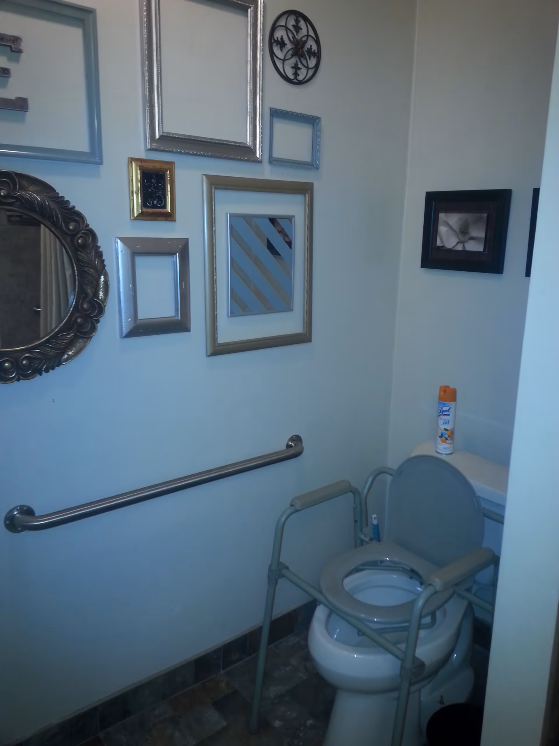 A bathroom with a toilet equipped with a raised seat and arm supports for accessibility. There is a grab bar mounted on the wall beside the toilet. The wall is decorated with various empty picture frames and a round ornate mirror. A can of Lysol air freshener is placed on top of the toilet tank.