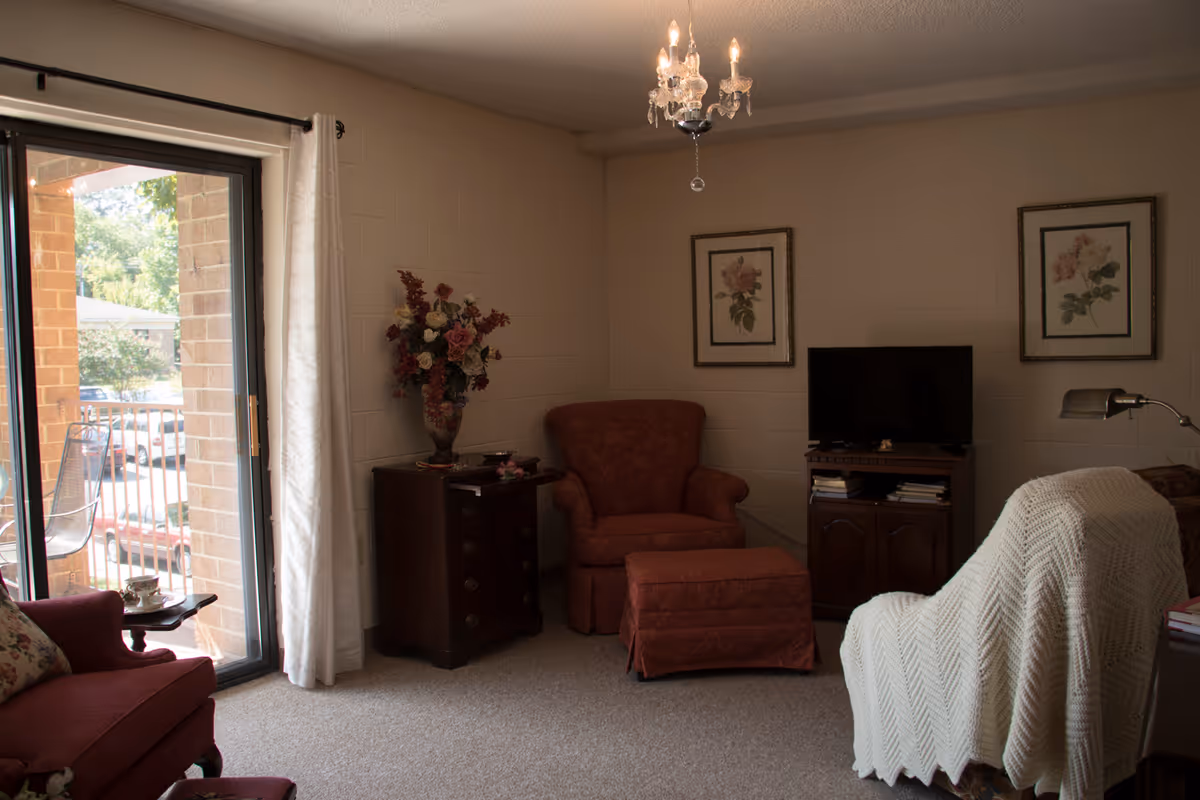 A cozy living room with a red armchair and matching ottoman, a wooden side table with a vase of flowers, a TV on a wooden stand, two framed floral pictures on the wall, a white knitted throw draped over another chair, and a sliding glass door leading to an outdoor balcony.