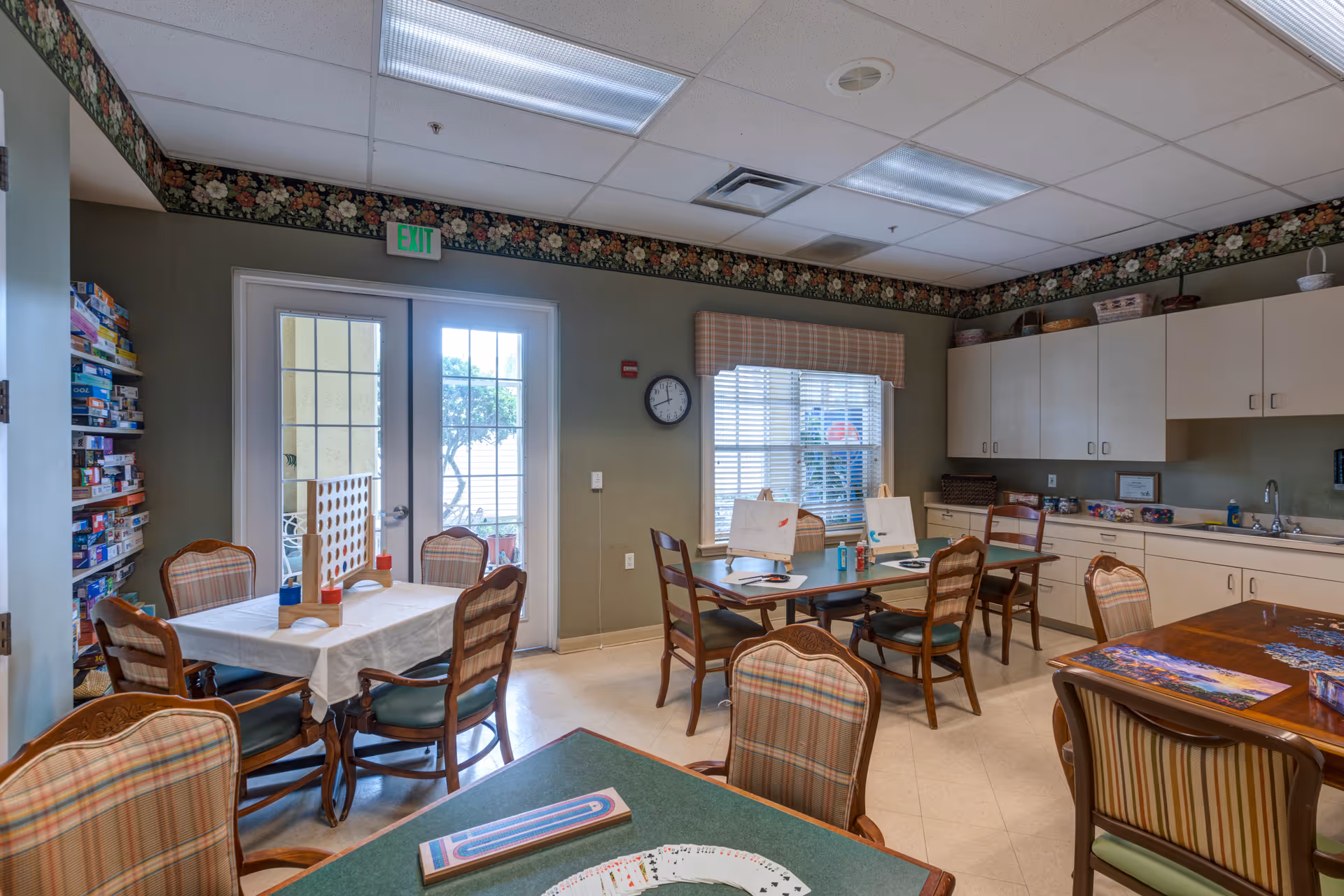 A well-lit activity room with several tables and chairs arranged for games and socializing. One table has a cribbage board and playing cards, another has a large Connect Four game, and shelves filled with board games are visible on the left. The room has a window with blinds, a clock on the wall, and cabinets with a sink along the right side. Floral wallpaper borders the ceiling.