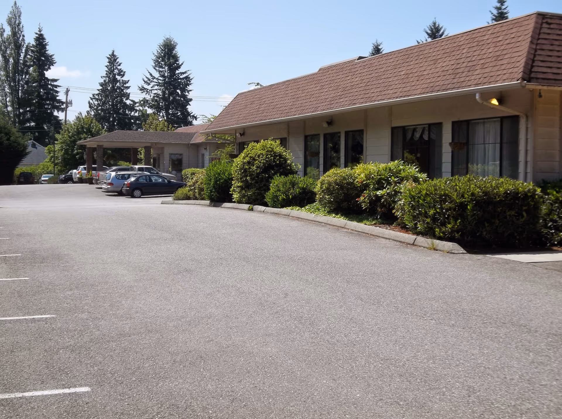 Exterior view of Arlington Health & Rehabilitation facility showing a single-story building with a brown shingled roof, several windows, and a covered entrance. There are bushes and greenery along the building and a parking lot with several parked cars in front. Tall trees and a clear blue sky are visible in the background.