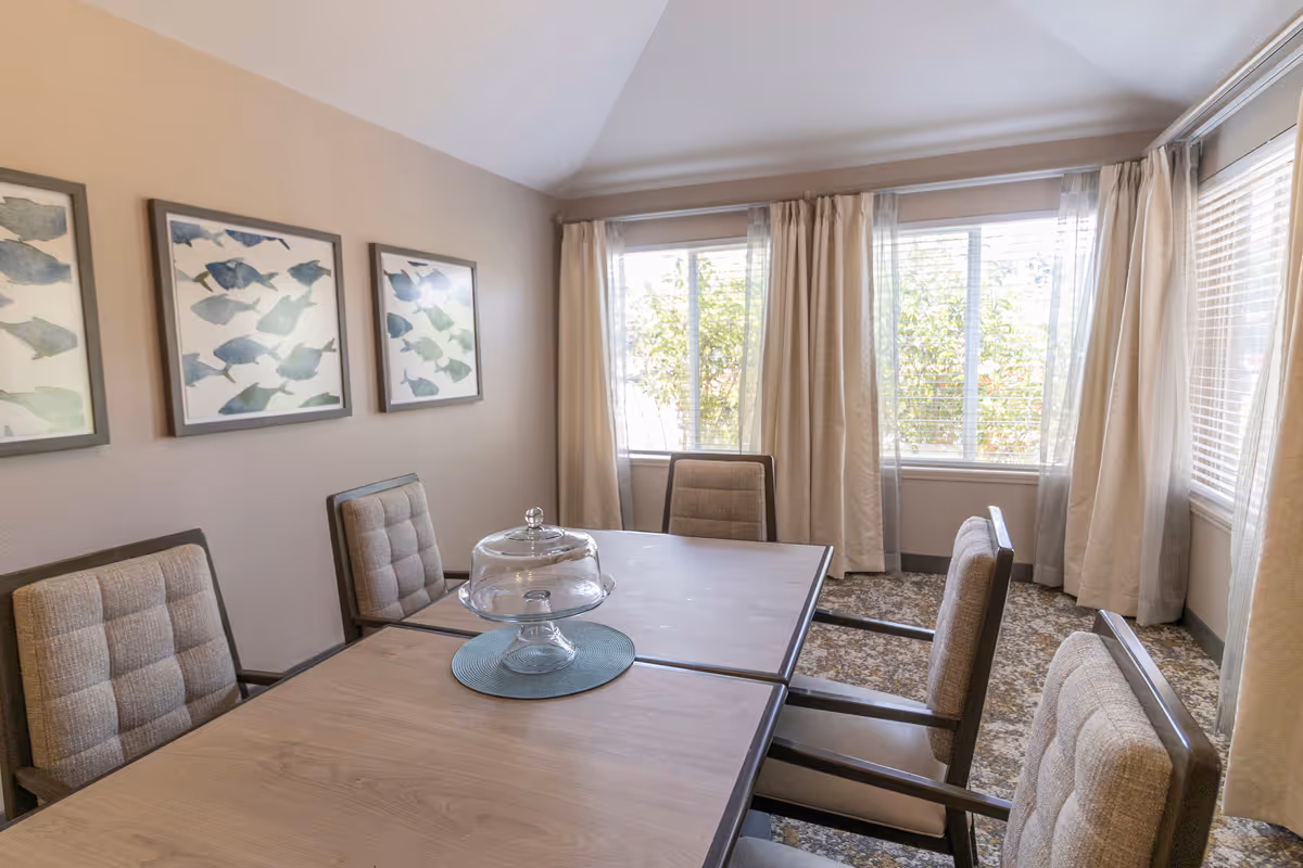 A small dining room with a wooden table surrounded by six cushioned chairs. The table has a glass cake stand with a lid placed on a blue placemat. The room has beige walls with three framed pictures of fish on one wall and large windows with beige curtains letting in natural light.