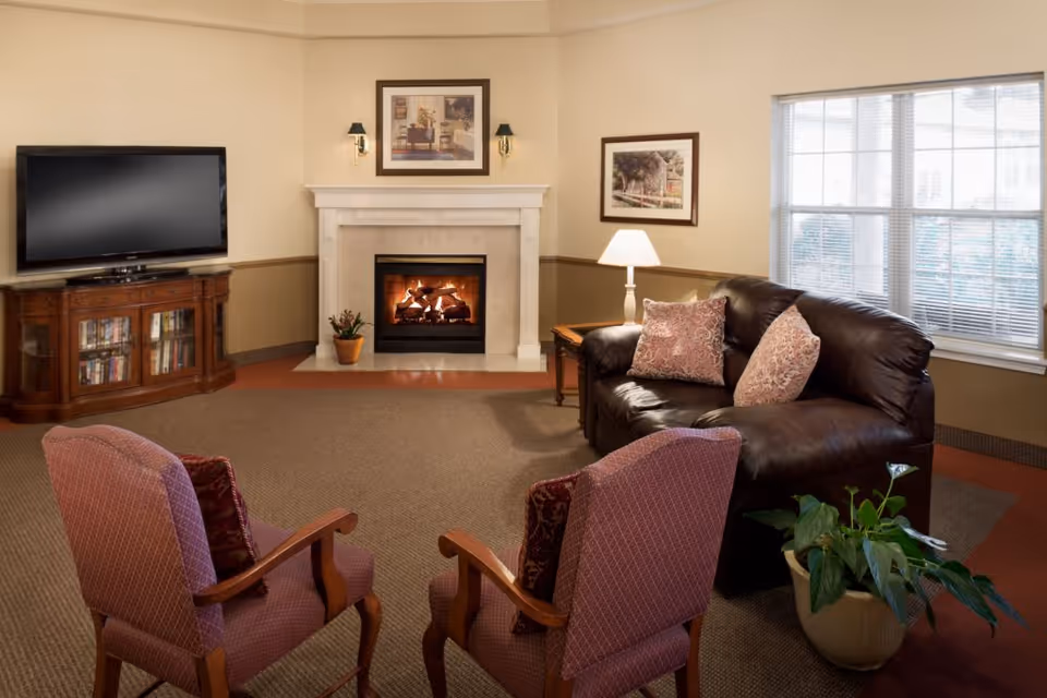 A cozy living room with a dark brown leather sofa adorned with two patterned pillows, two upholstered armchairs with wooden armrests, a wooden TV stand with a flat-screen television, a lit fireplace with a potted plant on the hearth, a side table with a lamp, framed artwork on the walls, a large window with blinds, and a potted plant on the floor.