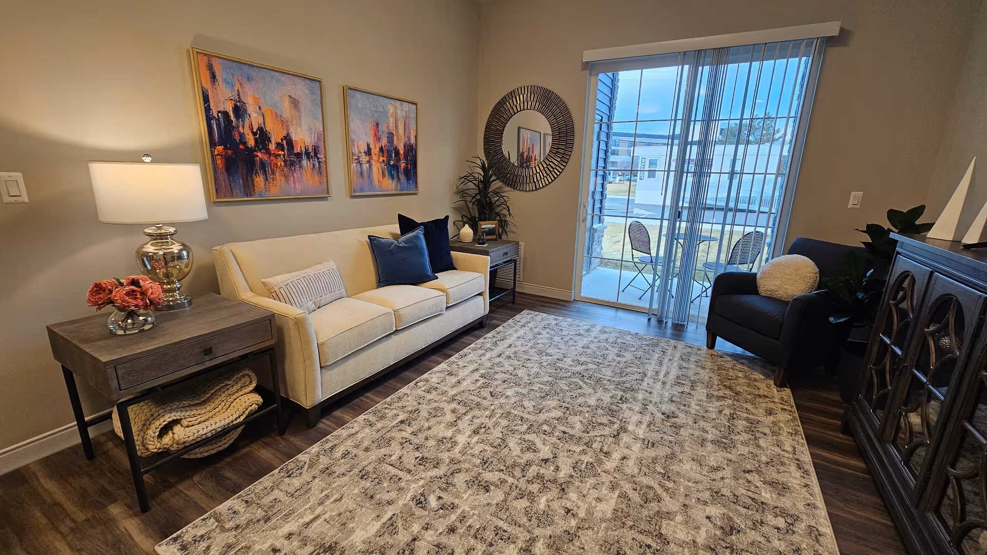 Well-decorated living room with a beige sofa, side tables, armchair, patterned rug, wall art and a sliding glass door opening to a small patio.