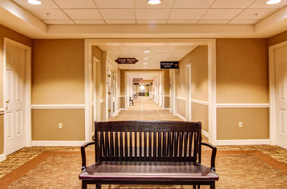 A long, well-lit hallway in a senior living facility with beige walls and carpeted floors. There is a dark wooden bench in the foreground. Several doors line both sides of the hallway, and signs hanging from the ceiling indicate rooms such as the Game Room and Veterans Room.
