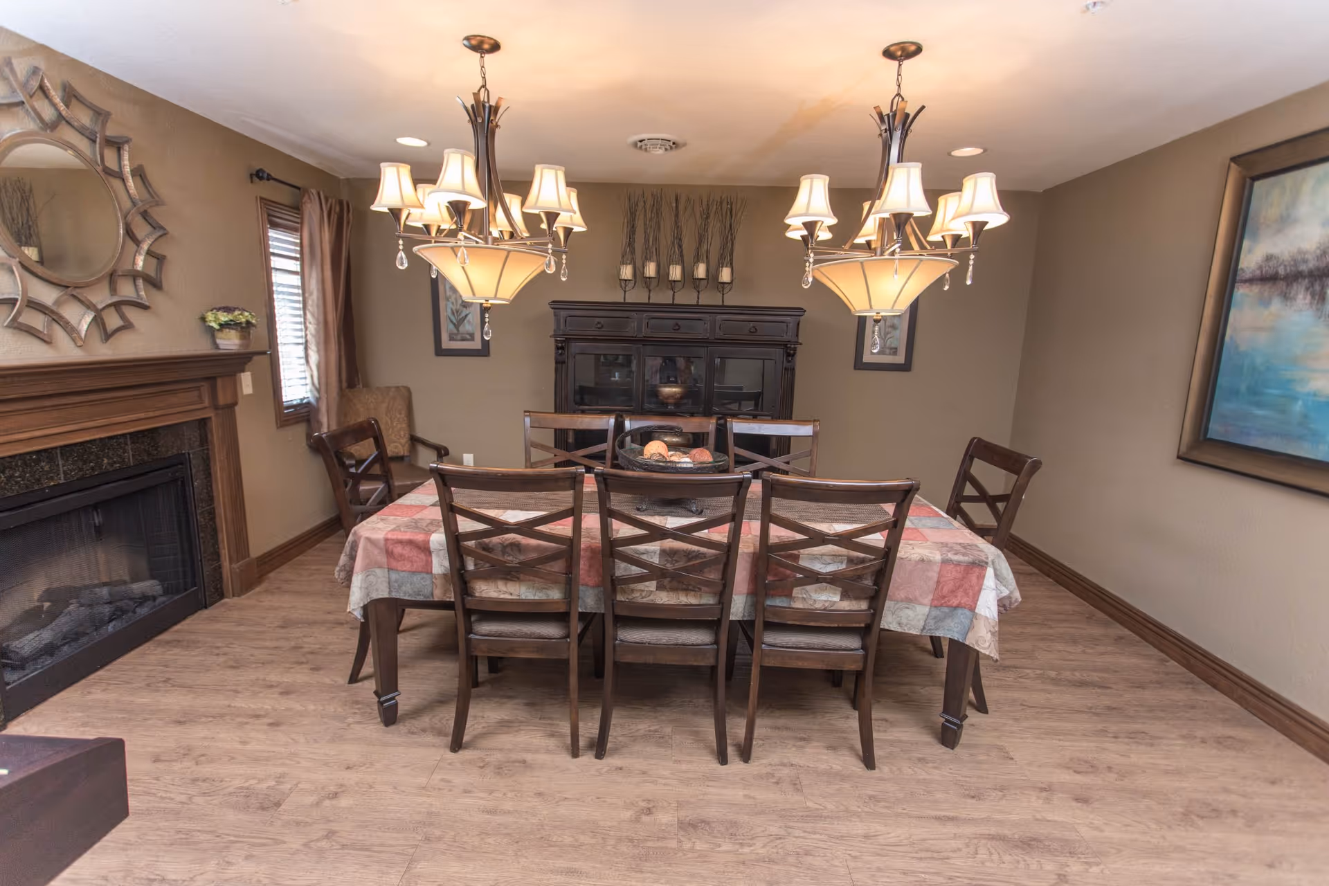 A dining room with a rectangular table covered by a patterned tablecloth and surrounded by eight wooden chairs. Two chandeliers with multiple lamps hang above the table. The room features a wooden floor, a fireplace with a decorative mirror above it on the left wall, a window with curtains, a dark wooden cabinet against the back wall, and framed artwork on the walls.