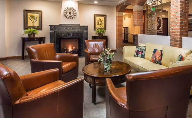 Comfortable seating area with four brown leather armchairs and a beige sofa around a round wooden coffee table in front of a lit fireplace and brick archways.