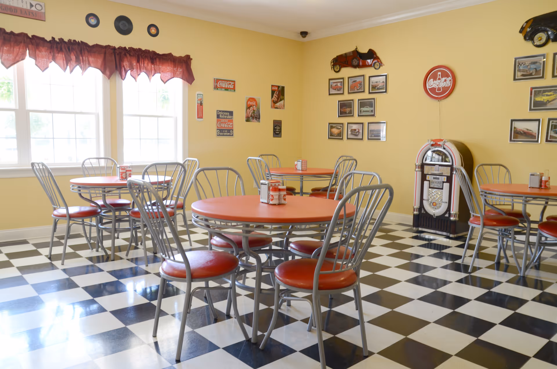 A retro-style dining room with black and white checkered floor, yellow walls, and red tables with matching red cushioned metal chairs. The room features large windows with red valances, vintage Coca-Cola signs and memorabilia on the walls, and a classic jukebox in the corner.