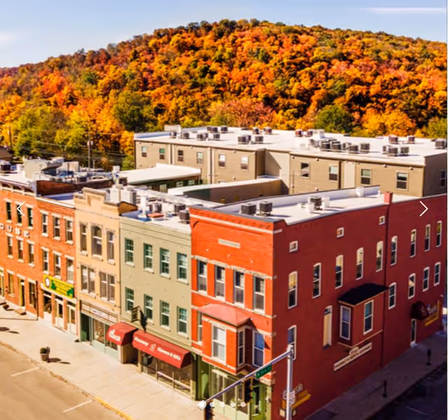 Aerial view of a small town street with multi-story brick buildings in front of a forested hill covered in autumn foliage with vibrant orange, yellow, and green colors.