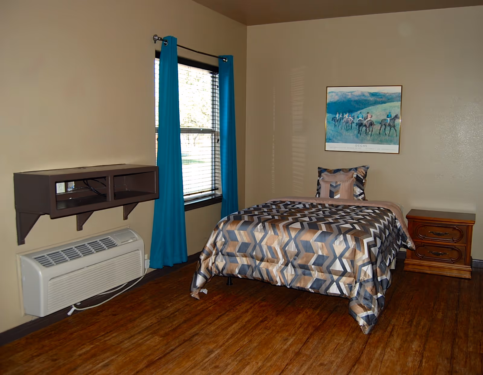 A simple single-occupancy bedroom with a patterned bed, nightstand, window with teal curtains, a wall-mounted shelf, and an air conditioning unit.