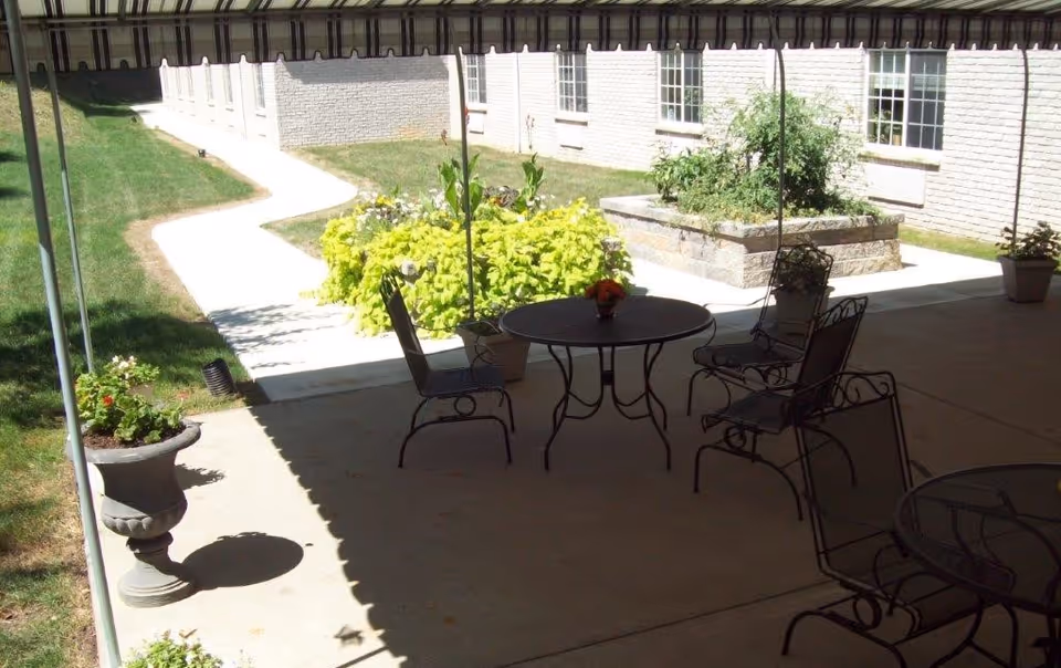 Outdoor patio area with metal tables and chairs under a striped awning. There are potted plants and a raised garden bed with greenery. A concrete pathway leads to a white brick building with windows.