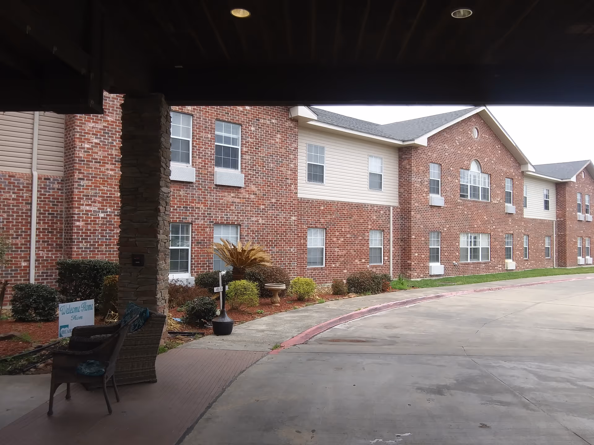Covered entrance and driveway in front of a two-story red brick senior living building with landscaping and seating.