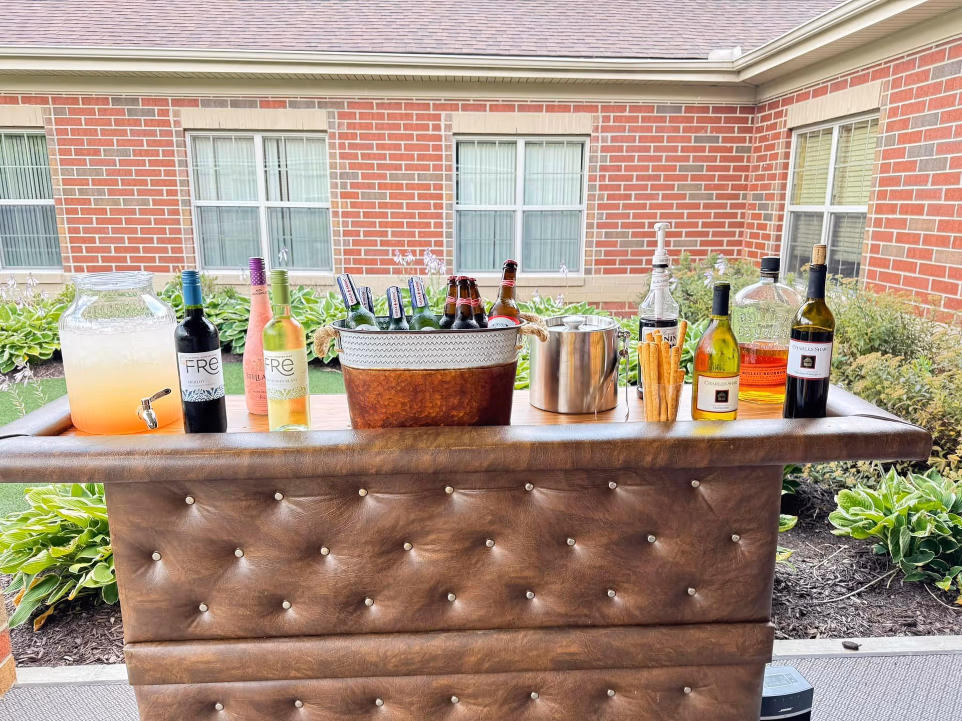 Outdoor bar setup with assorted wine, beer bottles, a beverage dispenser and ice bucket on a tufted brown bar in front of a brick building and windows.