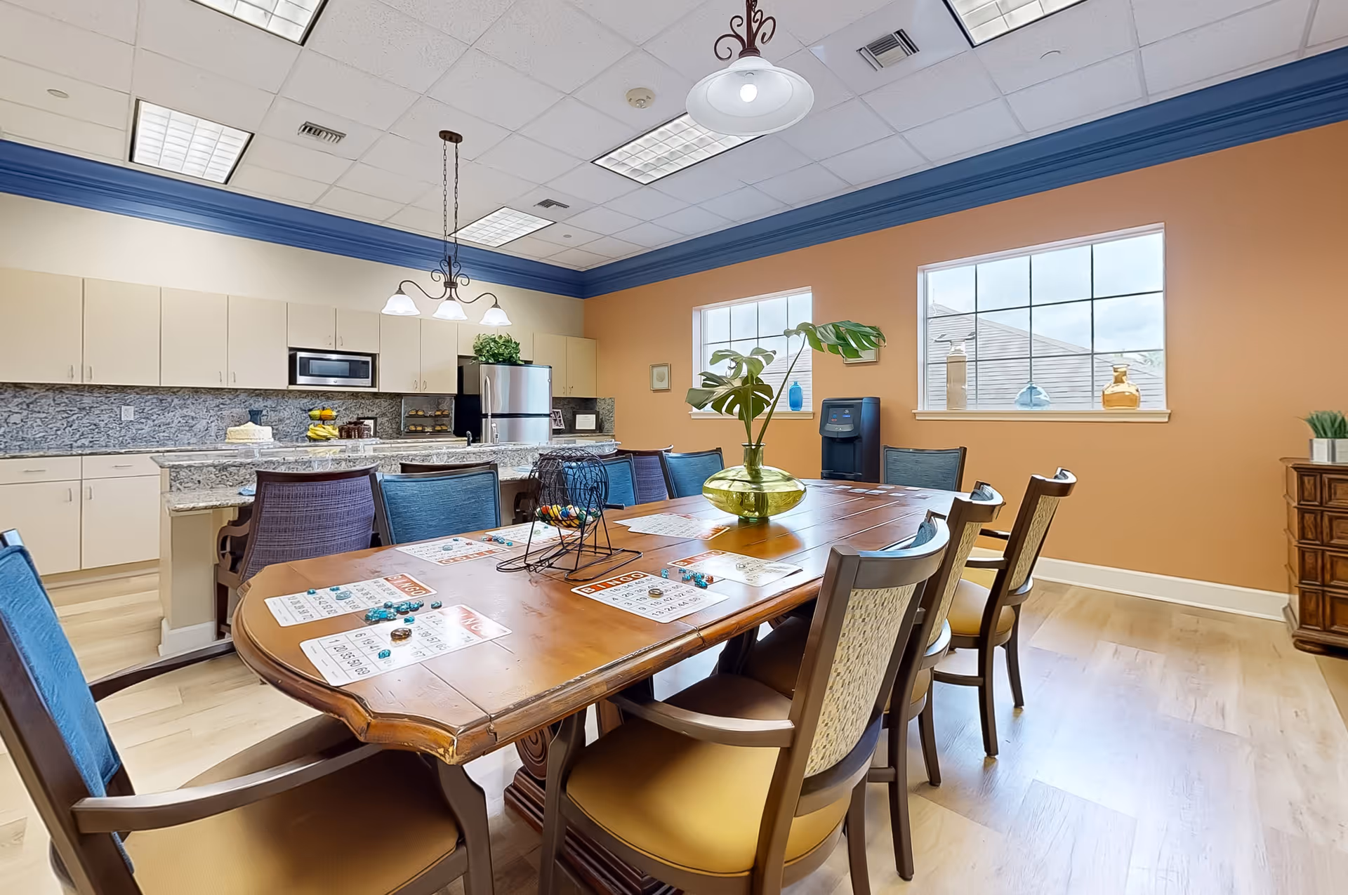 A bright and spacious room featuring a large wooden table with chairs arranged around it. The table has bingo cards and game pieces on it, indicating a recreational activity setup. Behind the table is a modern kitchen area with white cabinets, a granite countertop, a microwave, and a refrigerator. The walls are painted in a warm orange color with two windows letting in natural light. A decorative plant in a green vase is placed on the table, and a water dispenser is visible near the windows.