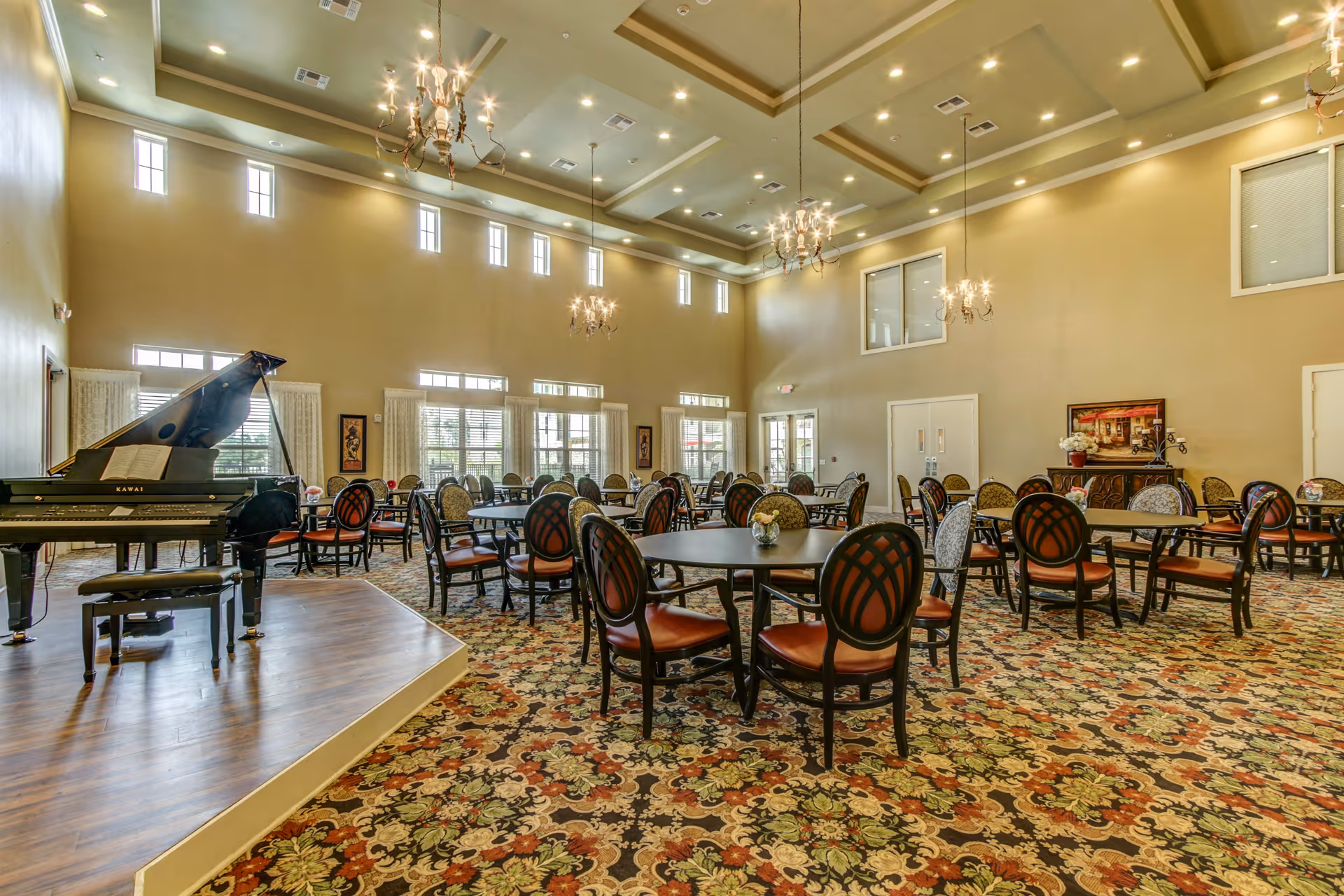 Large elegant dining/activity room with round tables and chairs, chandeliers, and a grand piano on a small stage.