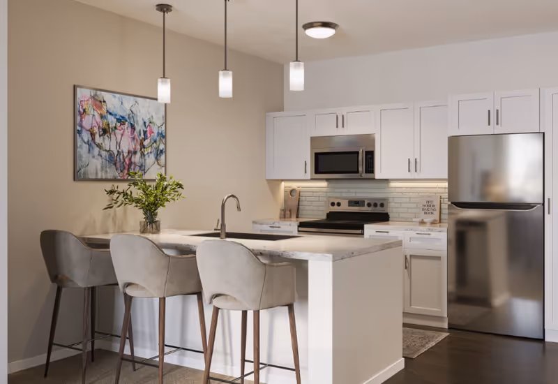 Modern kitchen with white cabinets, stainless steel refrigerator and microwave, an electric stove, and a kitchen island with a sink and three beige bar stools. A colorful abstract painting hangs on the wall, and three pendant lights hang above the island.