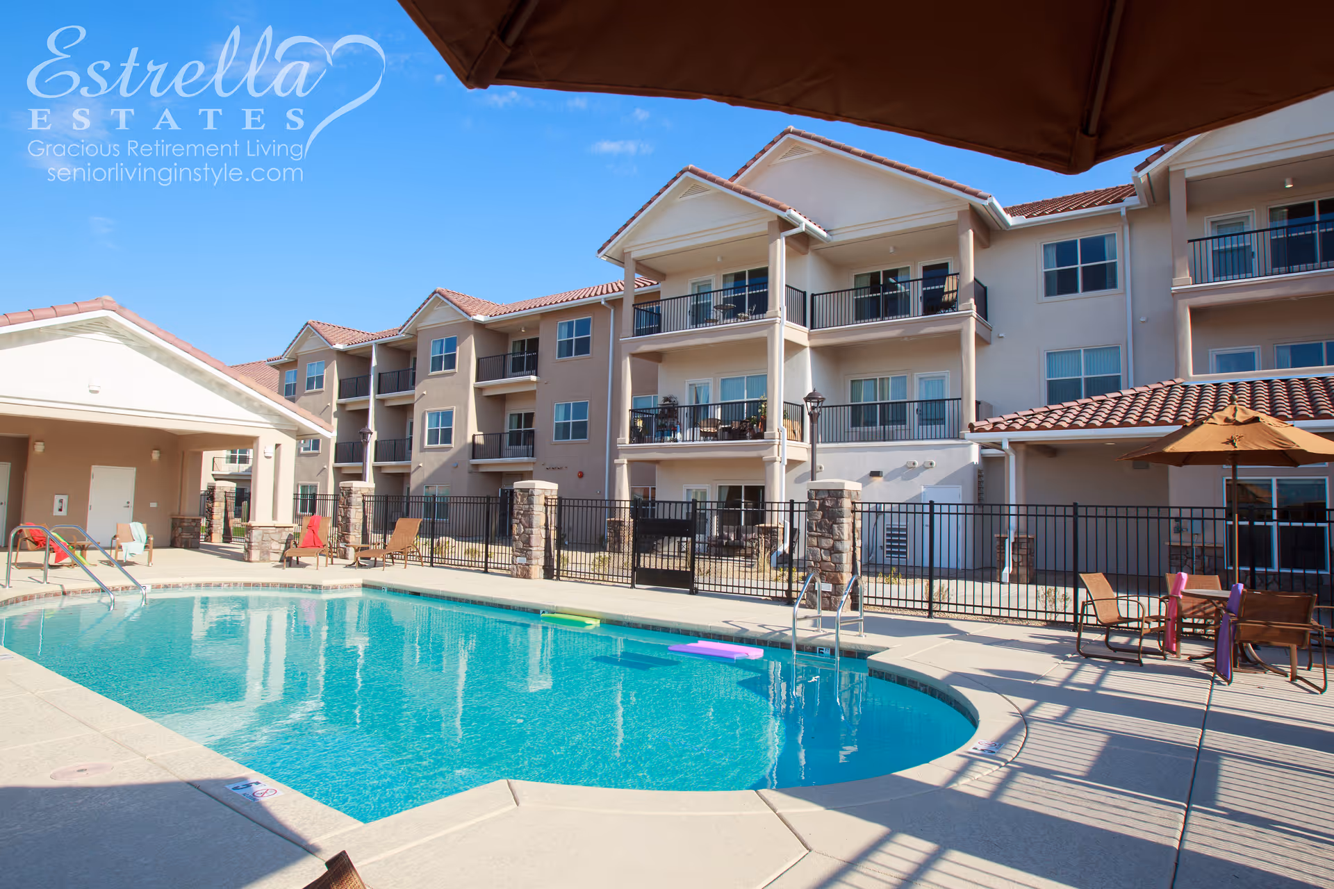 Outdoor swimming pool and patio with lounge chairs and umbrellas in front of a multi-story retirement community building with balconies.