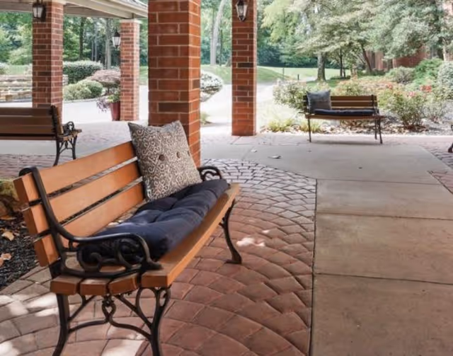 Outdoor covered seating area with wooden benches featuring decorative cushions, brick pillars, and a paved walkway surrounded by greenery and trees.