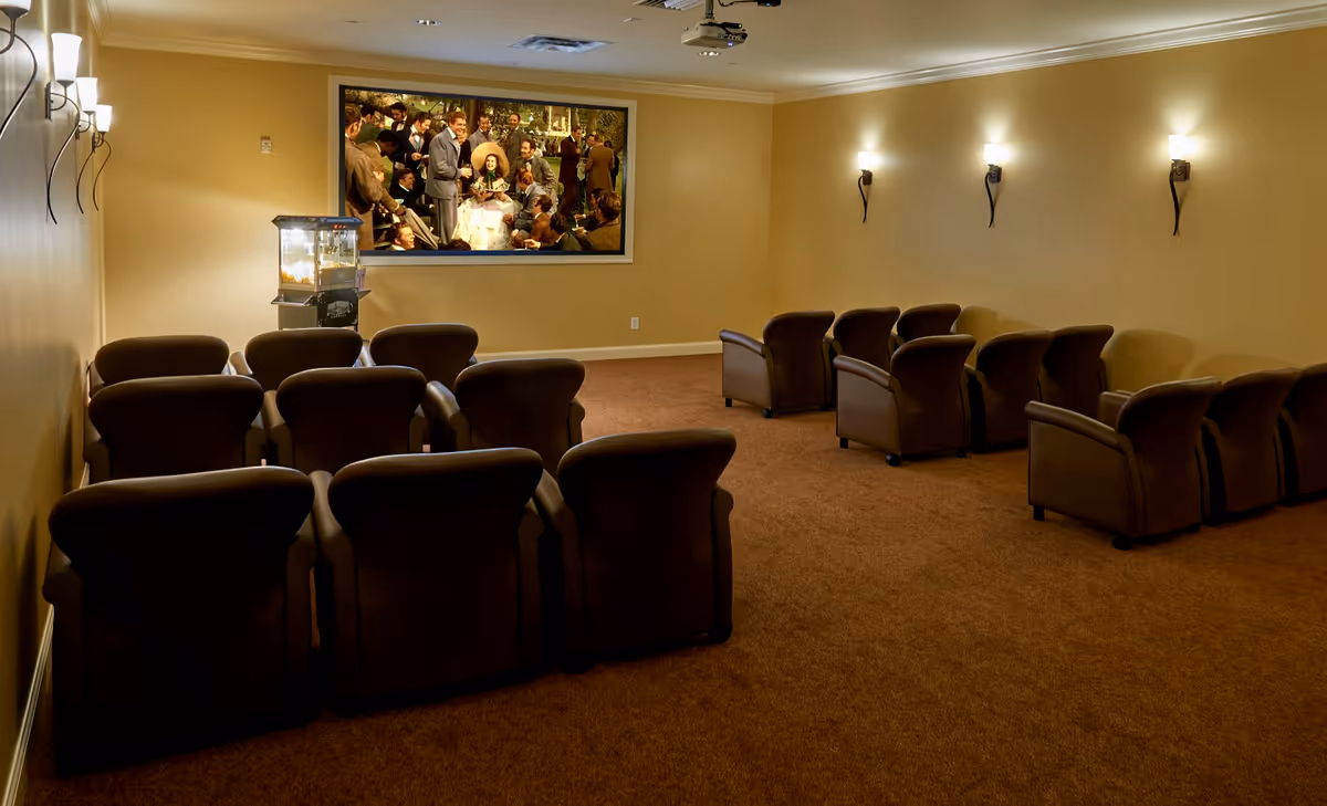 A small theater room with three rows of brown leather recliner chairs facing a large screen showing a movie scene. The room has beige walls, brown carpet, wall-mounted lights, and a popcorn machine near the screen.