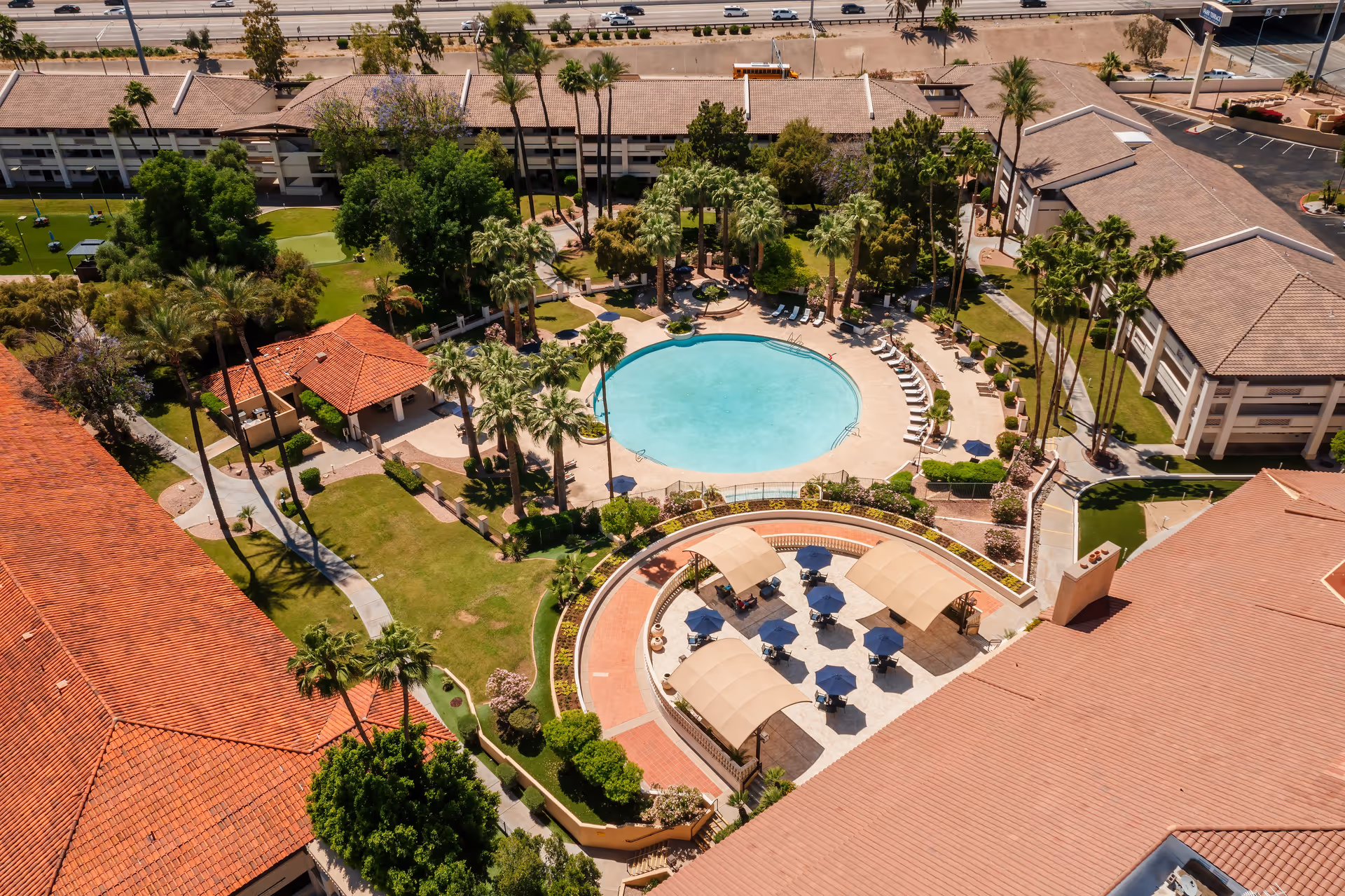 Aerial view of an outdoor area at a senior living facility featuring a circular swimming pool surrounded by lounge chairs and palm trees. There are shaded seating areas with tables and umbrellas nearby, well-maintained lawns, walking paths, and multiple buildings with red-tiled roofs surrounding the courtyard.
