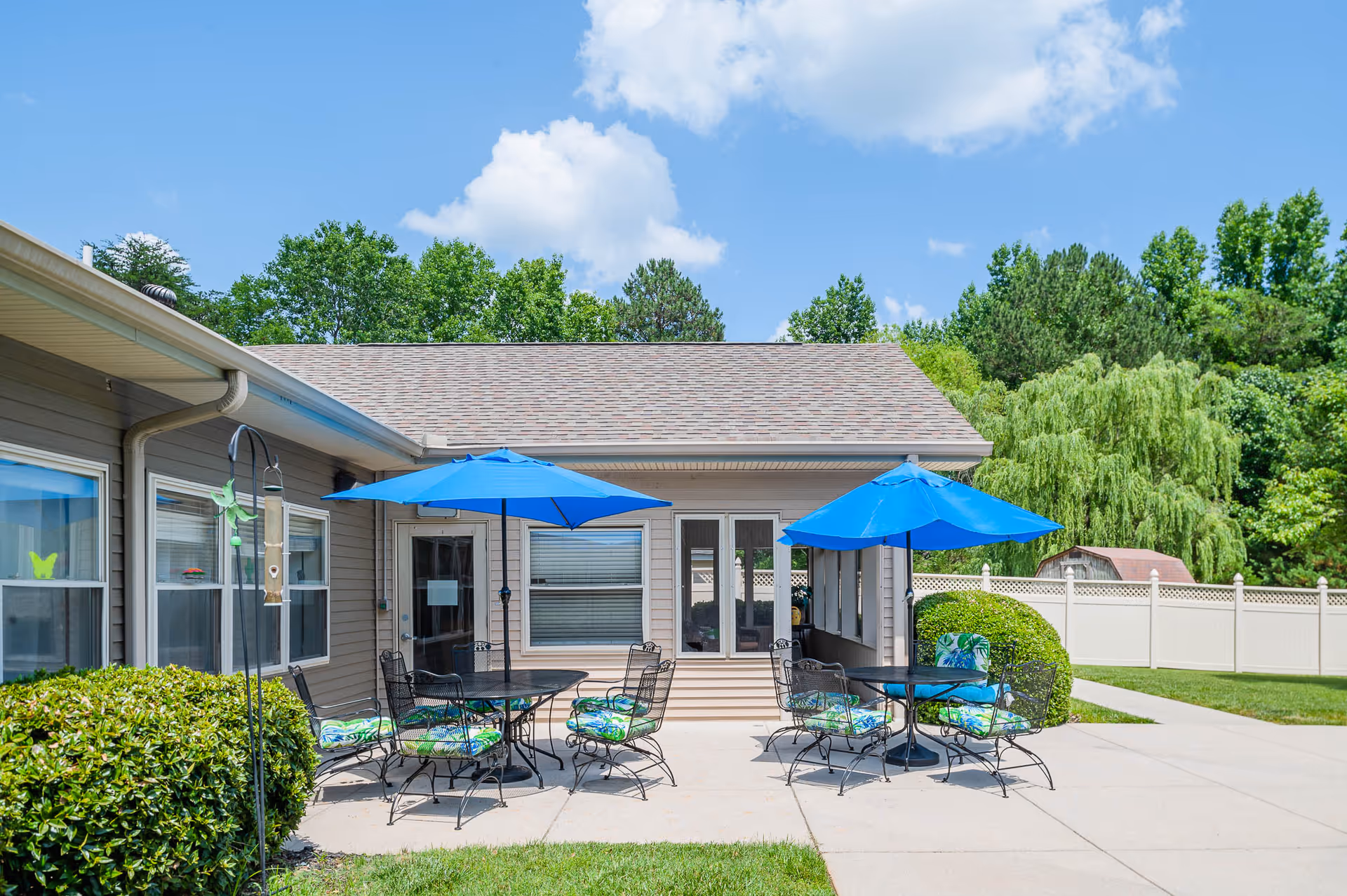 Outdoor patio area at Ascension Living Valley Residence with two round tables, each shaded by a blue umbrella and surrounded by metal chairs with colorful cushions. The patio is adjacent to a beige building with windows and a door, and is bordered by green bushes and a white fence. Trees and a blue sky with clouds are visible in the background.