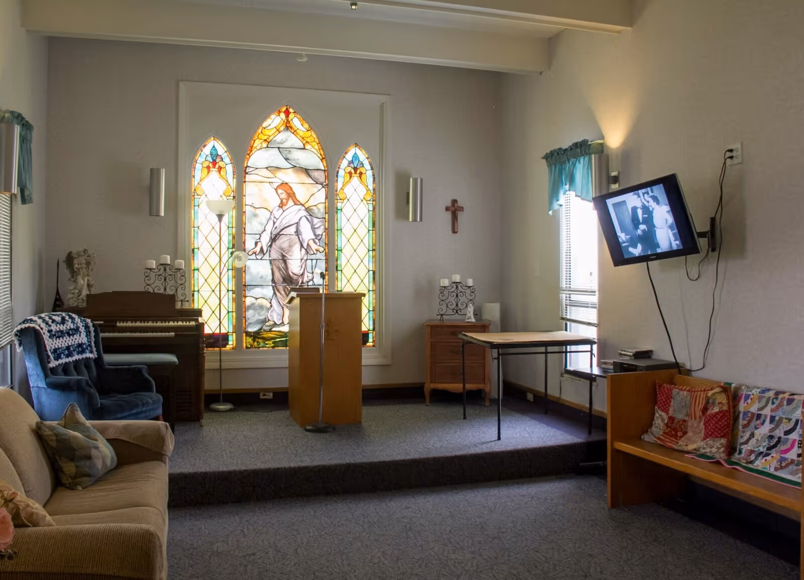 Interior of a small chapel or prayer room with a stained glass window depicting Jesus, a wooden podium with a microphone, a piano, a blue armchair with a crocheted blanket, a beige couch with pillows, a wooden bench with colorful quilts, a wall-mounted TV showing a black and white image, and a small wooden table and cabinet with candles and a cross on the wall.