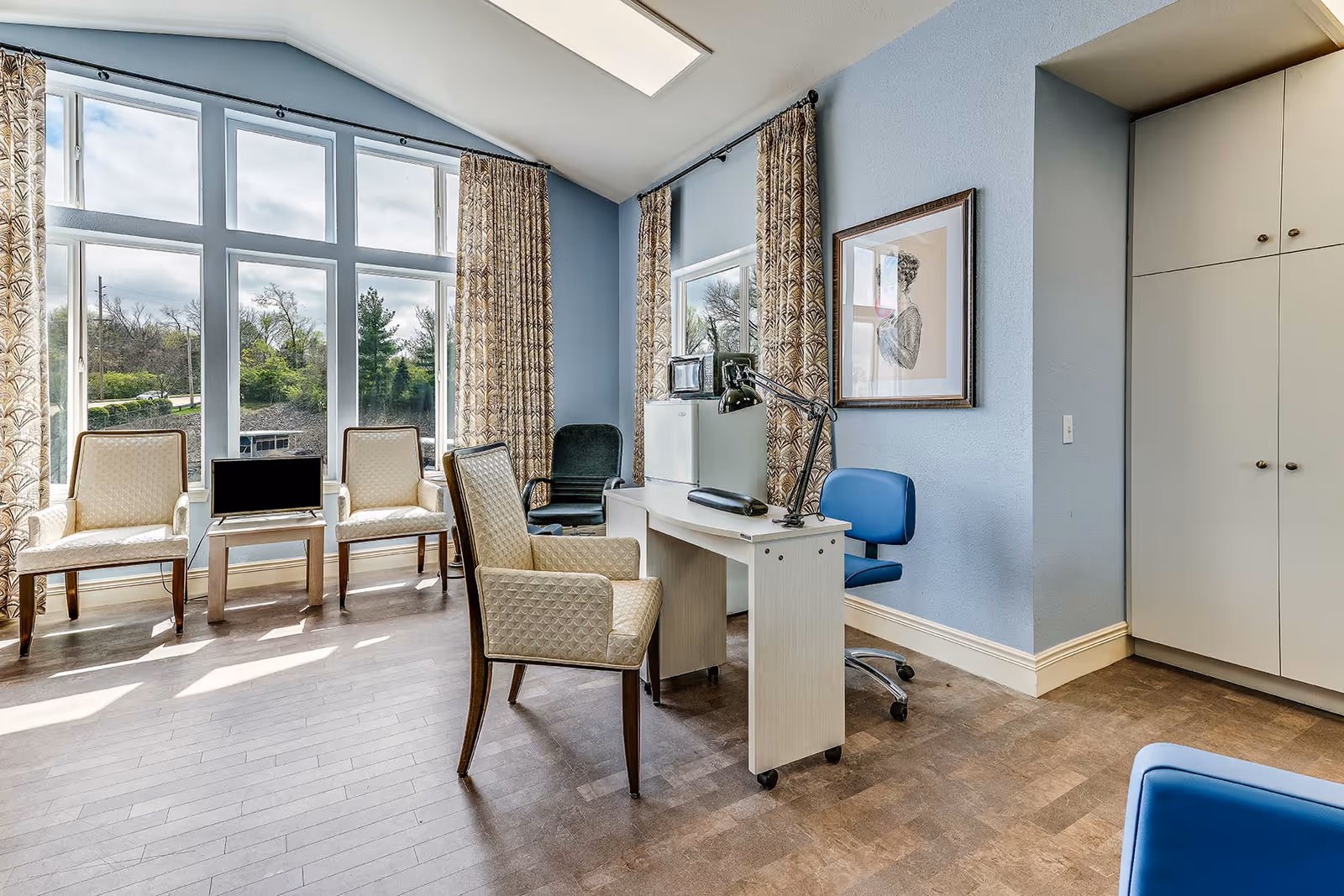 Bright senior living common room with large windows, several chairs, a desk with a computer and cabinetry.