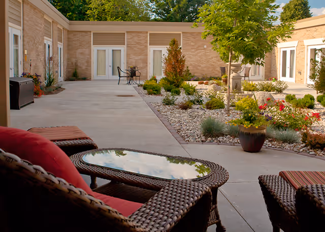 Wicker patio seating and glass-topped table overlooking a landscaped courtyard with potted plants, walkways, and ground-floor apartment doors.